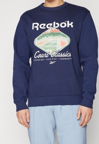 Marinblå sweatshirt med vitt Reebok-logotyp, tennisbana grafik och texten "Court Classics, Motion, Fair Play, Gemenskap," bärs av en person.