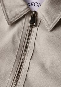 Lys beige jakke med en struktureret krave, subtil struktur og en metallynlås, der løber ned ad fronten. Mærketiketten er synlig indeni.