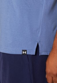 Gros plan d'une personne portant une chemise bleue avec une fente latérale et une étiquette noire avec le logo Under Armour au-dessus d'un pantalon bleu marine.