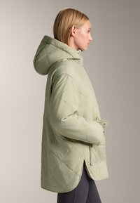 Žena v svetlo zelenej prešívanej bunde s kapucňou a prednými vreckami, stojaca v profile na neutrálnom pozadí.
