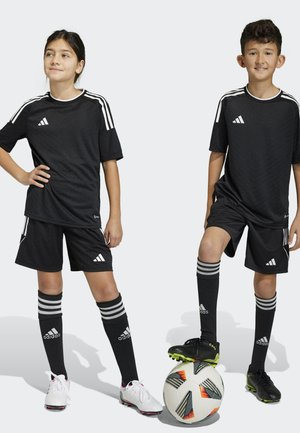 Schwarze adidas Sporttrikots und Shorts mit weißen Streifen, kombiniert mit schwarz-weißen Fußballschuhen. Ein gemusterter Fußball liegt auf dem Boden.