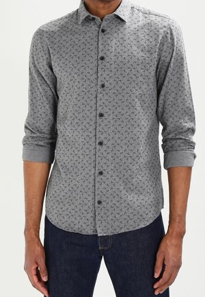 Mężczyzna ubrany w szarą koszulę na guziki z długim rękawem ozdobioną małymi czarnymi wzorami liści oraz ciemnoniebieskie dżinsy, z rękawami podwiniętymi do przedramion.