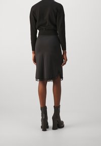 Kvinna iklädd en svart tröja, knälång svart kjol med spetskant och svarta högklackade ankelstövlar, stående med ryggen vänd.