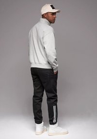 Jasnoszara bluza z wysokim kołnierzem, czarne spodnie z białymi paskami po bokach, białe sneakersy za kostkę oraz kremowa czapka bejsbolowa.