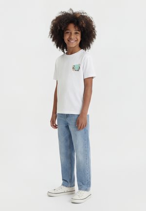 Bambino sorridente con capelli ricci che indossa una t-shirt bianca con grafica, jeans azzurri e scarpe da ginnastica bianche, in piedi davanti a uno sfondo semplice.