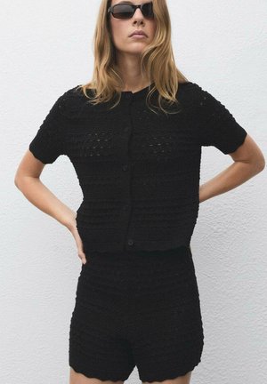 Ensemble en crochet noir composé d'un haut à manches courtes avec des boutons et d'un short assorti, tous deux ornés de motifs texturés et de bords festonnés.