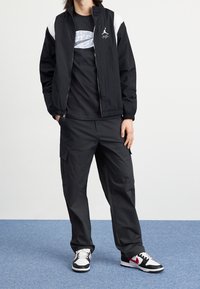 Černá lehká bunda s bílými akcenty, černé tričko s logem a černé cargo kalhoty. Obuv má černo-červený design.