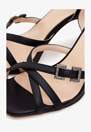 Sandalias negras de tacón alto con correas de cuero entrelazadas y una hebilla metálica cuadrada adornada con strass en el lateral. Plantilla suave de color claro.