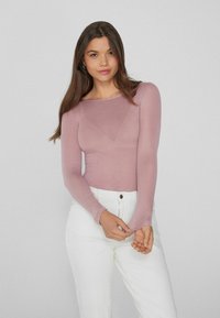 Langsleeve top in zacht pastelroze stof met een aansluitend ontwerp en een bootnek. Gecombineerd met witte jeans met hoge taille.