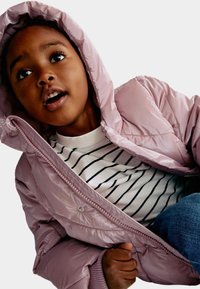 Chaqueta de plumón rosa claro con capucha, con cierre de cremallera en la parte frontal y broches a presión, llevada sobre una camiseta blanca de rayas y jeans azules.