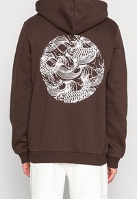 Bruine hoodie met een witte grafiek van draaiende vissen op de achterkant. Gemaakt van zachte stof met een trekkoordkap en geribbelde onderrand.