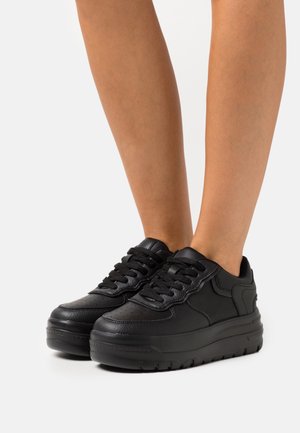 Benenen met zwarte leren platformsneakers met dikke zolen en zwarte veters, staand op een witte ondergrond.
