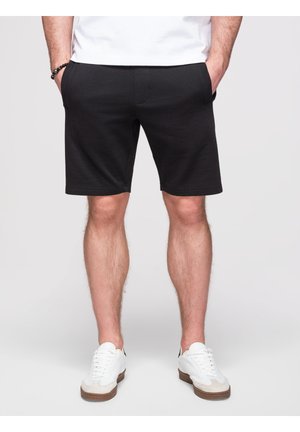 Mann trägt schwarze knielange Shorts, weiße Sneaker und ein weißes Hemd, mit den Händen in den Taschen, stehend auf einem schlichten hellen Hintergrund.