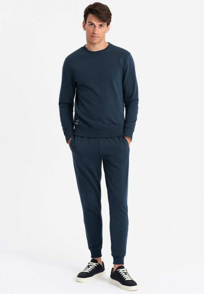 Sudadera azul marino con puños y dobladillo de canalé, combinada con pantalones ajustados a juego. El conjunto presenta un sutil logotipo de la marca en la cinturilla.