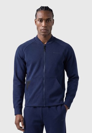 Man with braided hair wearing a dark blue zip-up jacket and matching pants, standing with one hand in pocket against plain background.
