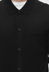 Cardigan noir boutonné avec col pointu porté sur un maillot blanc, avec une poche poitrine sur le côté gauche.
