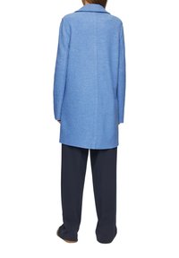 Cappotto lungo azzurro chiaro realizzato in tessuto strutturato, con colletto a risvolto, taglio dritto e dettaglio della cucitura posteriore. Abbinato a pantaloni a gamba larga.