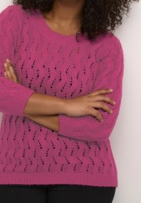 Person med krydsede arme iført en lyserød strikket sweater med et zigzag hulmønster mod en lys baggrund.