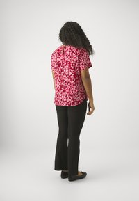 Red and pink patterned short-sleeve top, curved hem, paired with black flared pants and black flats, with curly hair visible from the back.