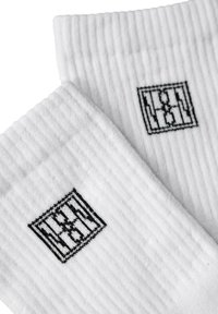 Białe prążkowane skarpety z czarnym geometrycznym logo na ściągaczu. Materiał jest miękki, a faktura gładka i elastyczna.