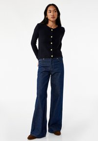 Navy neulecardigan, jossa on tekstuuri, suuret kultaiset napit. Yhdistetty leveälahkeisiin tummiin farkkuihin ja ruskeisiin loafereihin.
