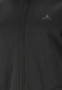 Svart zip-up jacka med textilstruktur, platta sömmar och en liten logotyp i övre vänstra hörnet. Slätt material med en figurnära passform.