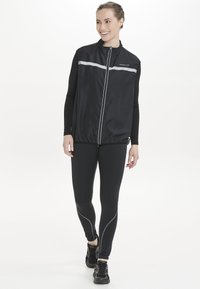 Svart lättviktsväst med reflexband, dragkedja och hög krage, i kombination med åtsittande svarta leggings och träningsskor.