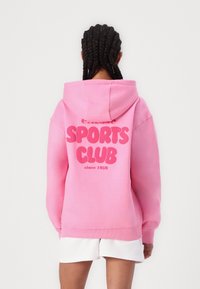 Personne aux cheveux tressés porte un sweat à capuche rose avec le texte « SPORTS CLUB since 1959 » dans le dos et un short blanc, debout devant un fond uni.