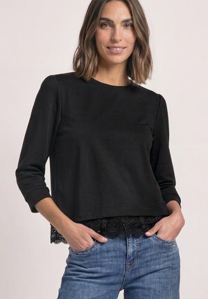 Femme aux cheveux mi-longs portant un haut noir à manches longues avec un ourlet en dentelle et un jean bleu, mains dans les poches, souriant légèrement.