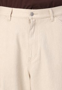 Beige cotton pants with a straight fit, featuring a button closure, zip fly, and side pockets. Fabric has a smooth texture and diagonal weave.