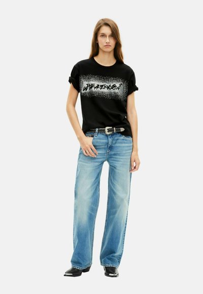 Vrouw draagt een zwarte T-shirt met opgerolde mouwen en witte grafische tekst, lichtblauwe wijde jeans, een gesp met studs en zwarte puntige schoenen, staand tegen een witte achtergrond.