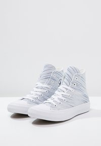 Bílé vysoké tenisky s modrým pruhovaným vzorem, s bílými šňůrkami, gumovou špičkou a logem značky na boku.