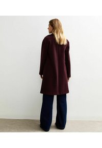 Lungo cappotto bordeaux con un design su misura, caratterizzato da un taglio dritto, maniche lunghe e una ventrale sul retro, abbinato a jeans scuri a gamba larga.