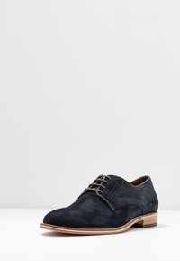 Chaussure lacée en suede marine avec bout arrondi, doublure en cuir marron et semelle contrastante en cuir marron clair. Détails de couture subtils sur les côtés.