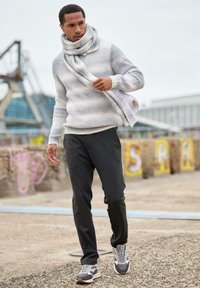 Hellgrauer gestrickter Pullover mit horizontalen Streifen, kombiniert mit schwarzen Hosen und grauen Turnschuhen. Ein passender Schal ist um den Hals gewickelt.