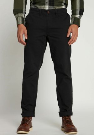 Homme portant un pantalon noir, une chemise à carreaux verte et blanche, et des bottes en cuir marron, debout les bras détendus le long du corps.