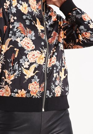 Musta vetoketjullinen takki, jossa vaaleanpunaisia ja keltaisia kukka- sekä kurkiprinttejä, päällä mustat housut, käsi koskettaa päätä.