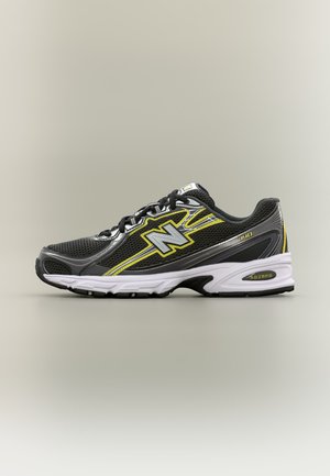 Zapatilla de correr New Balance negra y gris con detalles amarillos, cordones negros y suela blanca sobre un fondo beige liso.