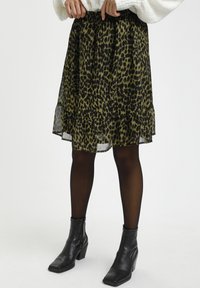 Nainen, joka on pukeutunut keskipitkään leopardikuvioiseen hameeseen, mustiin sukkahousuihin, mustiin korkokenkiin nilkkakorkoihin ja valkoiseen villapaitaan, säätää vyötärönauhaansa.