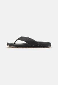 Mustat flip-flop sandaalit, joissa on teksturoitu hihna, pehmustettu pohja ja ruskea kumipohja, jossa on sahalaitakuviota. Brändin nimi on näkyvissä.