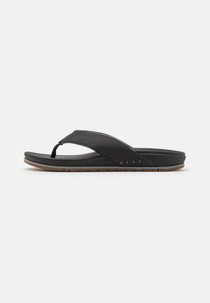 Black flip-flop sandal, featuring a textured strap, cushioned sole, and a brown rubber outsole with a zigzag pattern. Brand name is visible.