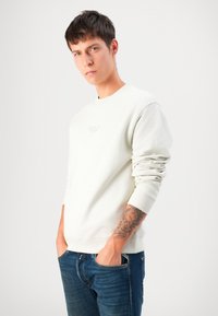 Young man with tattooed forearm wearing a white long-sleeve sweatshirt and blue jeans, standing with hands in pockets against white background.