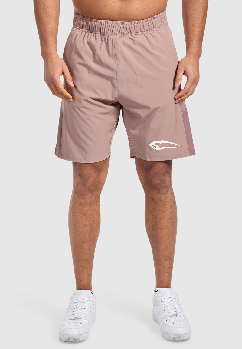 Homme portant un short de sport mauve clair avec un logo blanc sur la jambe gauche et des baskets blanches, debout devant un fond uni.