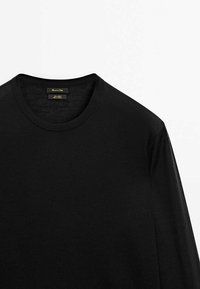 T-shirt noir à col rond et manches longues en tissu tricoté de qualité, présentant le haut de l'avant et le col avec l'étiquette de la marque à l'intérieur du col.