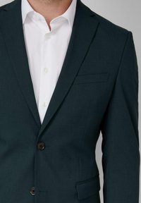 Chaqueta de traje en un verde oscuro con solapas con muesca, un bolsillo en el pecho y dos botones negros. Llevada sobre una camisa blanca de botones.