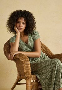 Mujer con cabello rizado vestida con un vestido verde estampado, sentada pensativamente en una silla de mimbre tejida contra un fondo liso beige.