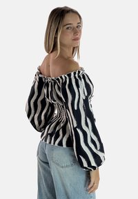 Schulterfreie Bluse mit schwarz-weißem Wellenmuster, gerafftem Ausschnitt und voluminösen Ärmeln, kombiniert mit hellblauen Jeans.