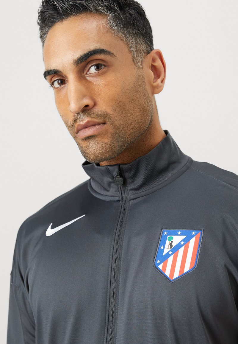 Veste de sport grise avec un col haut, logo Nike en blanc, et un blason coloré de l'Atlético de Madrid sur la poitrine gauche. Texture lisse.