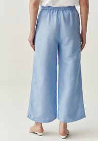 Lichtblauwe wijde broek met een elastische tailleband. De stof heeft een zachte textuur met een subtiele glans. De zoom is afgewerkt met een nette rand.