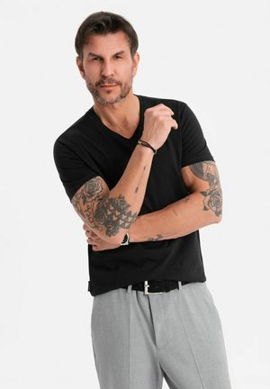 Camiseta negra de manga corta y escote en V combinada con pantalones estampados gris claros. Tatuajes visibles en los brazos. Cinturón negro con hebilla de plata.